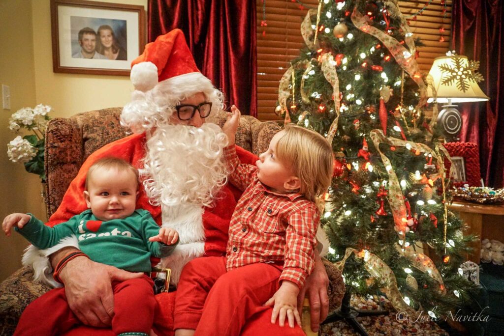 Image of two young children sitting on Santa's knee with a backdrop of a Christmas tree and holiday decor. Making the holidays about memories rather than gifts can help our children go on to lead more sustainable lifestyles.