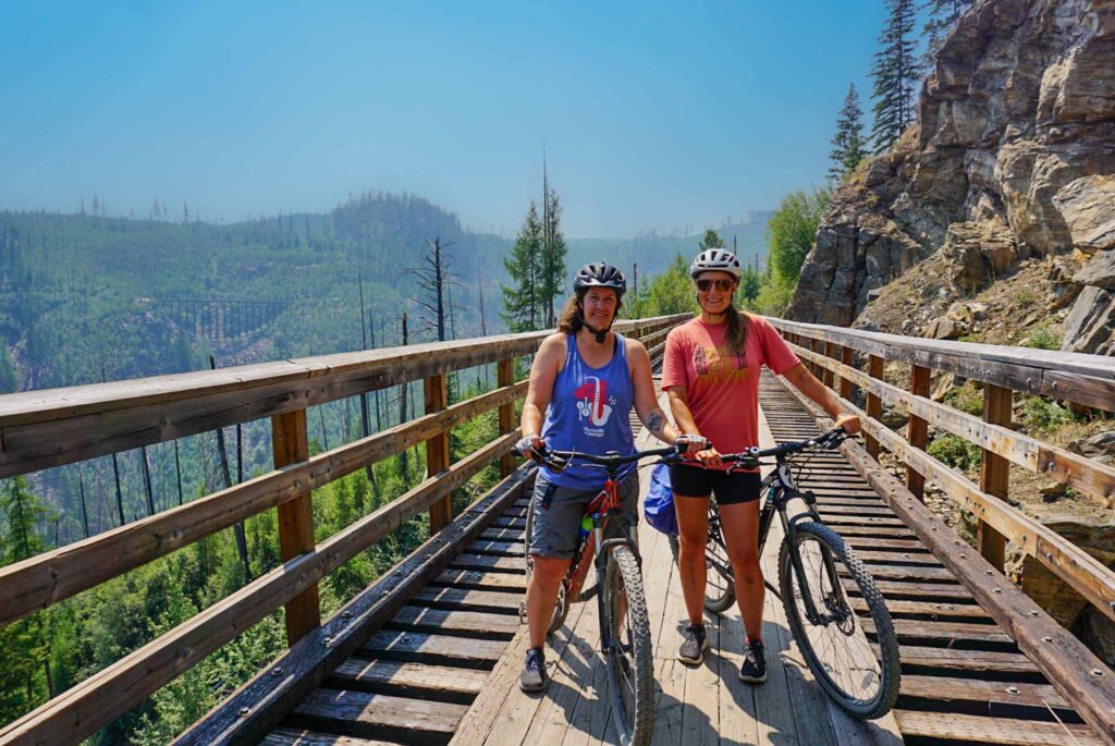 Image of two cyclists with their bikes on an old railway trellis with mountains in background. Choosing people-powered recreation can help you lead a more sustainable lifestyle.