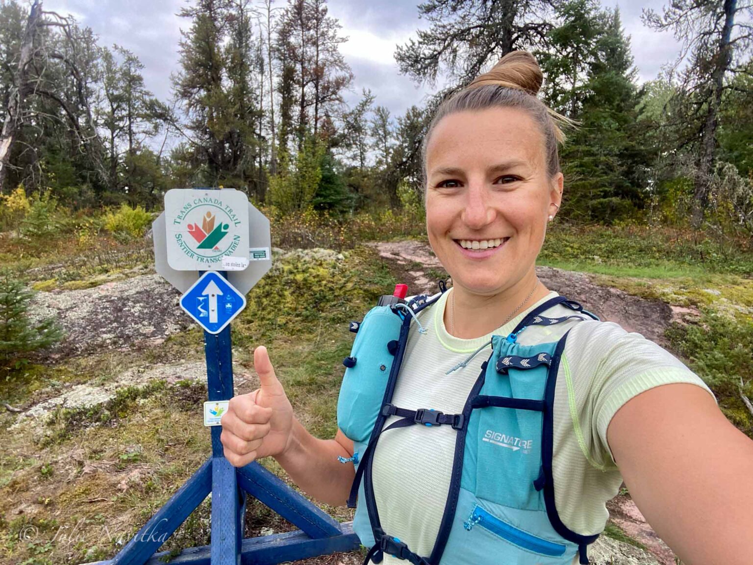 Image of a trail runner wearing a running vest gives a thumbs up next to a trail sign in Whiteshell Provincial Park, Manitoba, Canada. Choosing quality hiking and running gear is important when it comes to sustainable recreation.