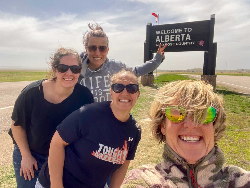 Image of four friends in front of the "Welcome to Alberta" sign on a windy day. Carpooling on a trip can make your travel method more sustainable.