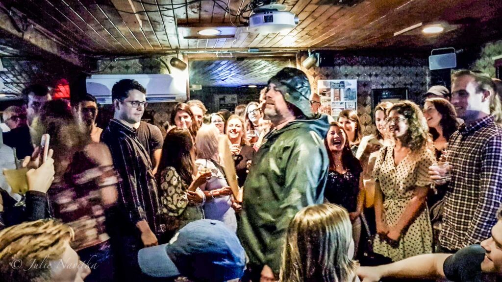 Image of a "screeching in ceremony" in a crowded bar room with man in fishing gear highlighted in centre. Taken in St. John's, Newfoundland, Canada. Learning about the local culture can help your city trip be more sustainable.