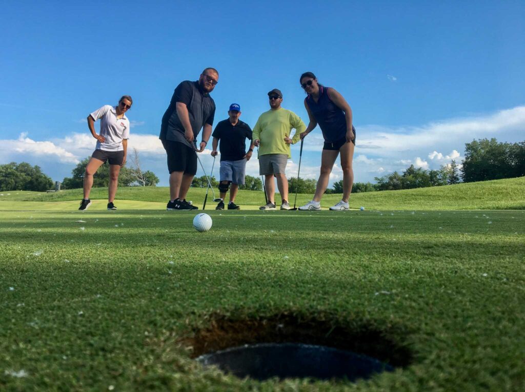 Image of five golfers with hole in immediate foreground.