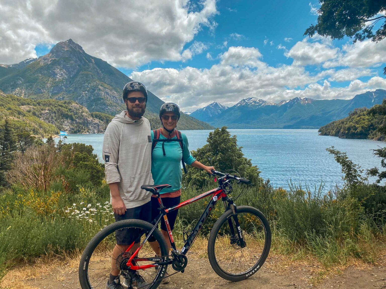 Young couple poses with mountain bike wearing helmets in front of picturesque background of lake and mountains. Human-powered is the most sustainable type of recreation.