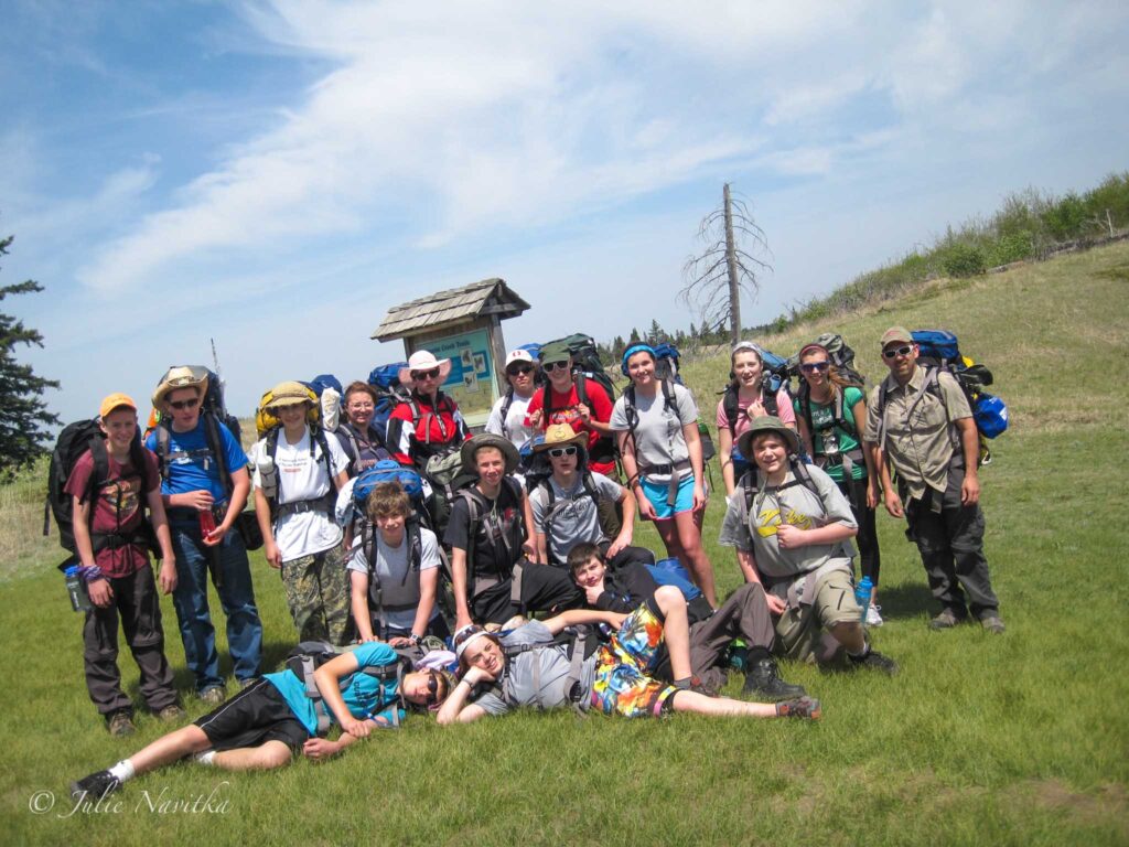Image of a school group of multi-day hikers wearing backpacks and hats in front of a trailhead sign. Spending meaningful time with children outdoors fosters a sustainable lifestyle.