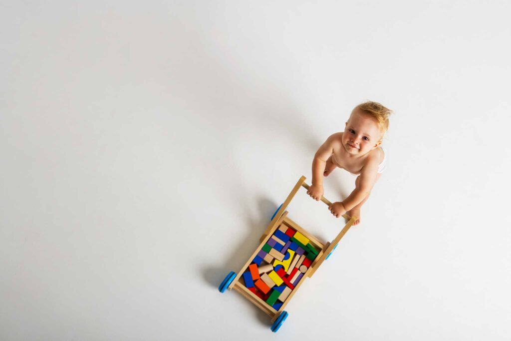 Birds eye image of a baby walking a wooden stroller filled with blocks with a white background. Choosing sustainable toys for children is essential for their health and environmental and social sustainability.