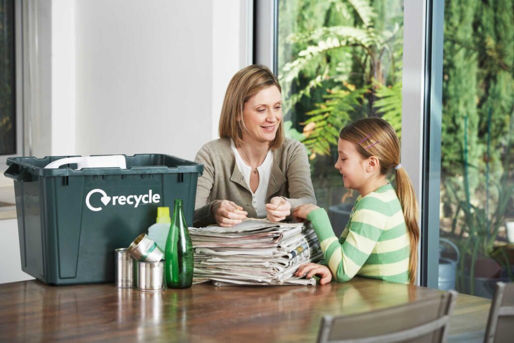 Image of a mother and child sitting at a table with a recycling bin and paper. Teaching our children about sustainability is essential.