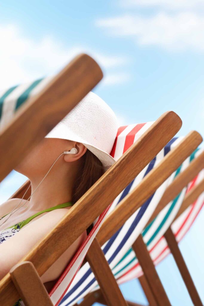 Cropped image of a beach goer wearing a hat relaxing on a sling-type beach chair in the sun. Wooden and organic cotton or linen chairs are great sustainable choices for a beach chair.