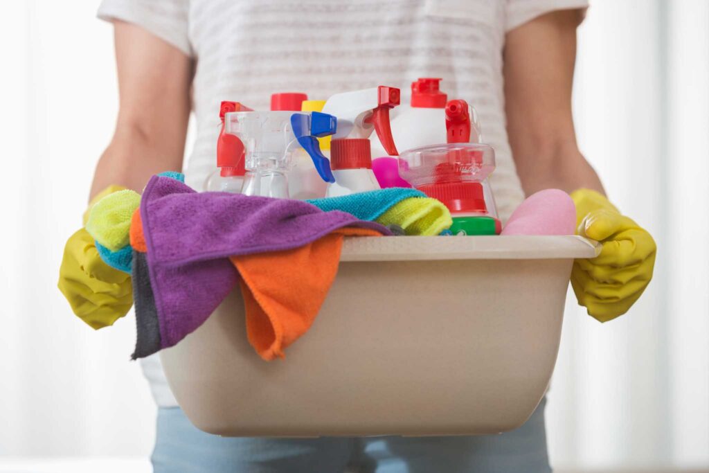 Close up image of a person wearing dish gloves holding a basin filled with cleaning products and clothes. Choosing eco-friendly cleaning products can help you create a more sustainable home.