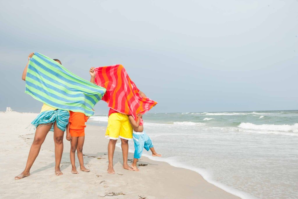 Image of a family of four hidden behind the brightly coloured beach towels they are waving around while standing at the water's edge on the beach. There are many sustainable materials from which beach towels and blankets can be made.