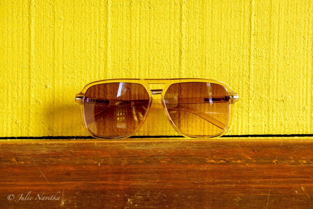 Image of a pair of sunglasses sitting atop a piece of wood in front of a bright yellow background wall. Choose sunglasses made ethically and from non-toxic materials for a sustainable lifestyle. 