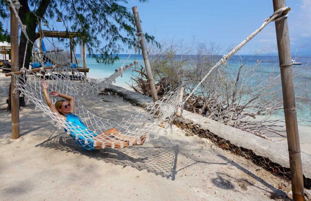 Image of a young beach-goer laying in a net hammock on a sunny day with a gorgeous beach and turquoise waters in the background. Eco-friendly beach products can help you practice sustainable recreation.