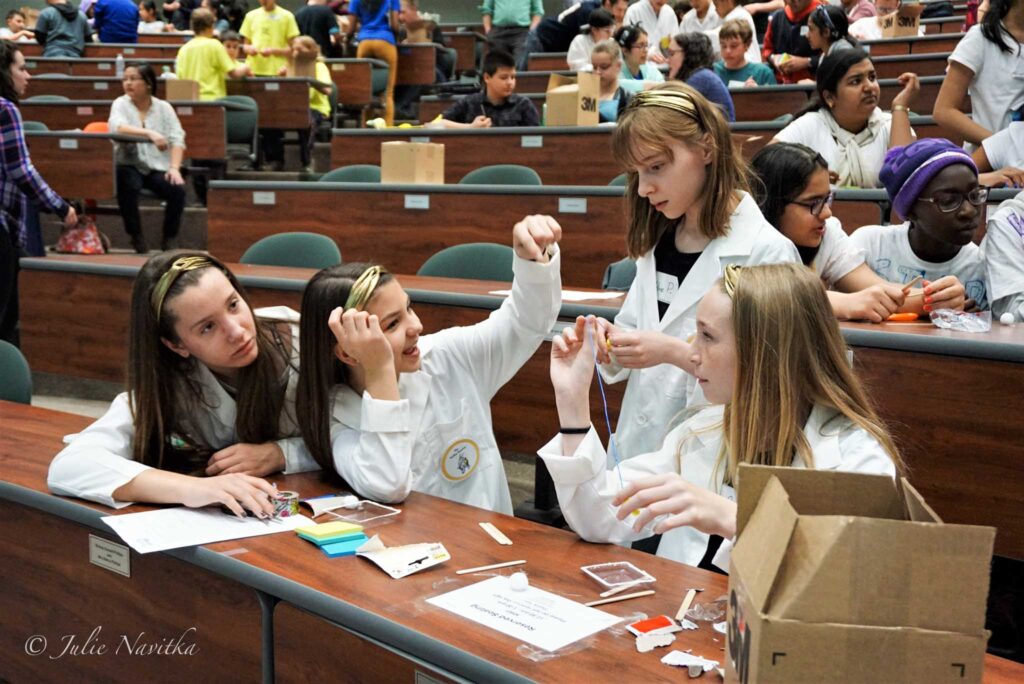 Image of four young students wearing lab coats in a lecture hall, collaborating on a project. Teaching about the environment and encouraging students to live a more eco-friendly lifestyle leads to a more sustainable school.