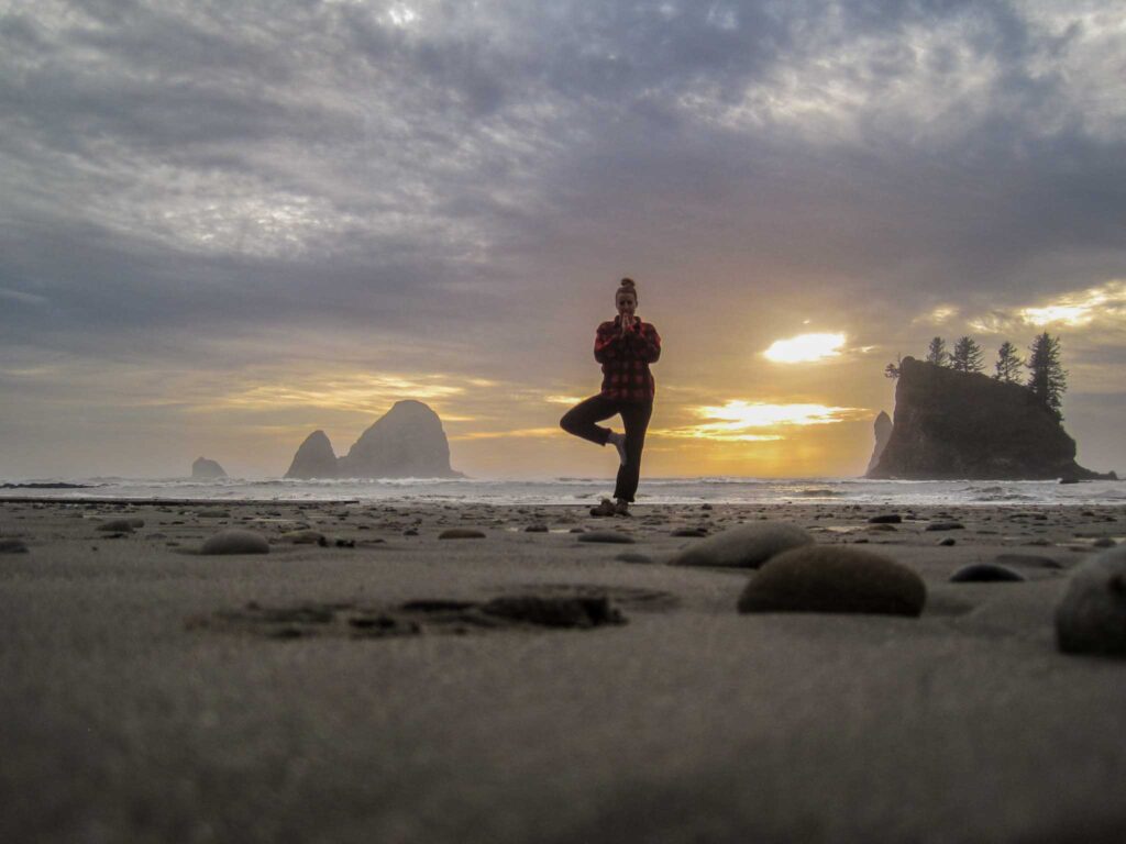Image of a silhouetted person standing in tree pose on a beach at sunset with rocky island formations jutting out of the water in the background in Olympic National Park, Washington State.