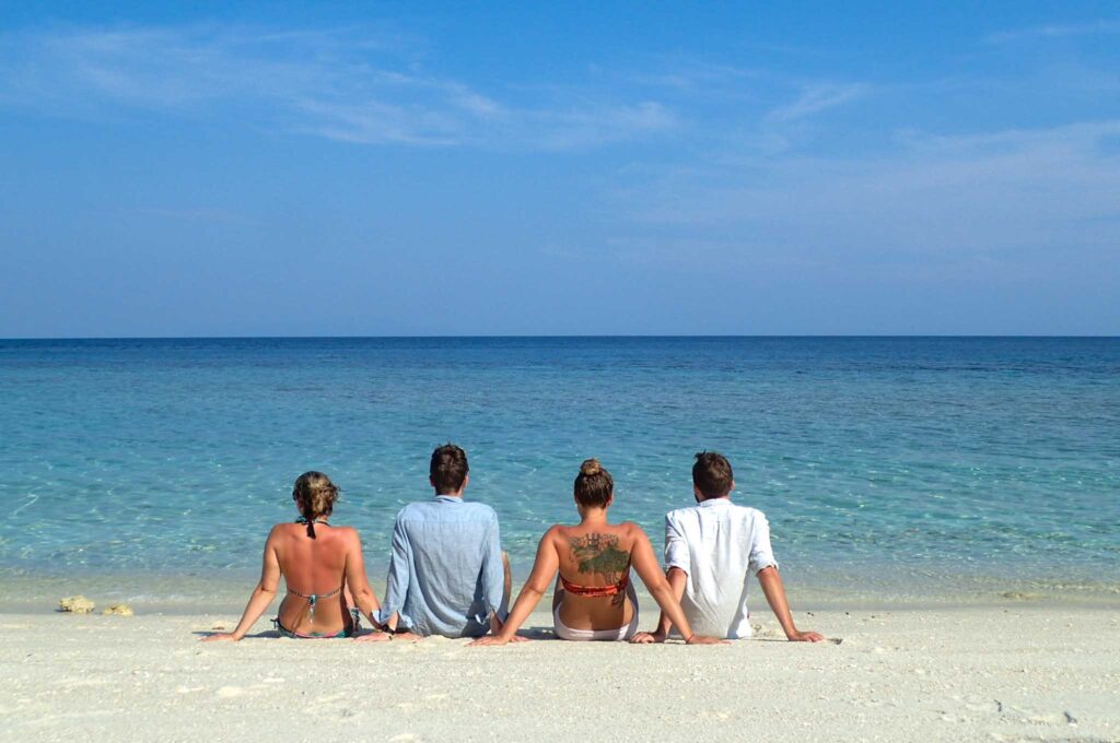 Four friends sitting side by side on a beach with backs to the camera with arms overlaced, looking out to a turquoise sea. Sustainability is possible at the beach.