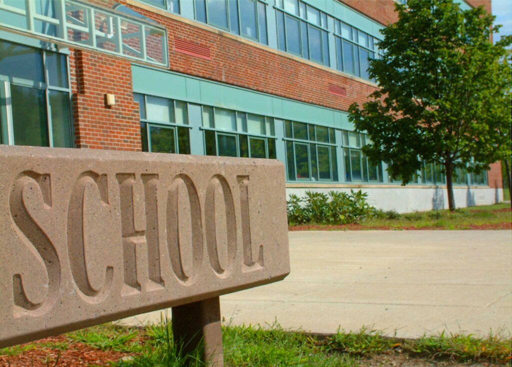 Close-up image of a sign reading "School" with a school building in the background. Making schools sustainable requires collaboration from all levels of the education system.