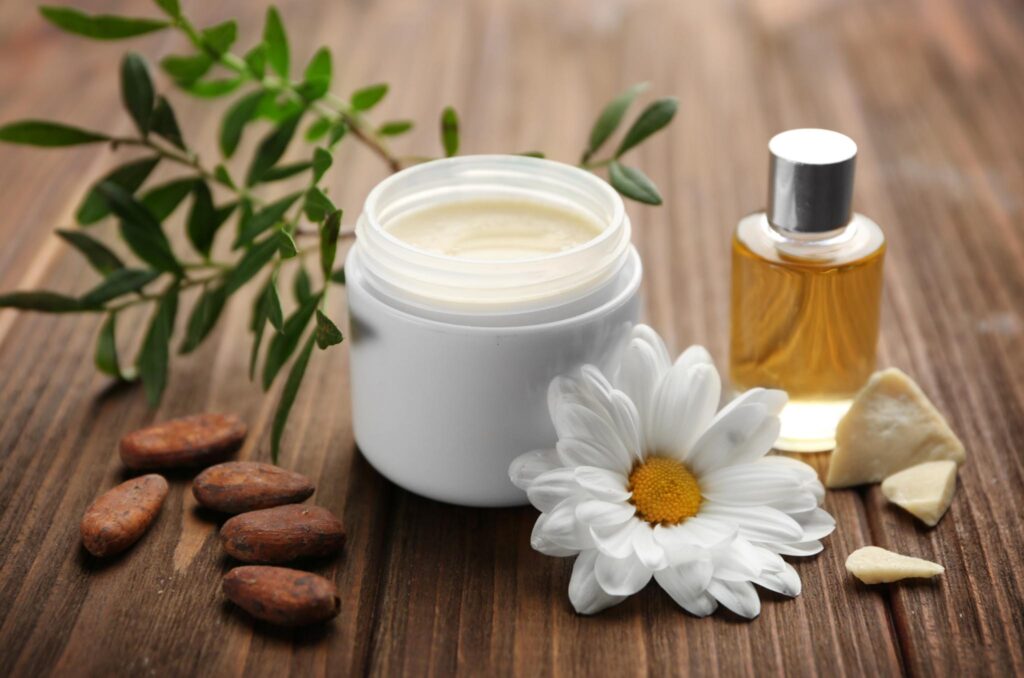 Image of a jar of white lotion amongst natural ingredients such as almonds, daisy flower, and essential oil. Creating your own beauty products can lead to a more sustainable health and beauty routine.