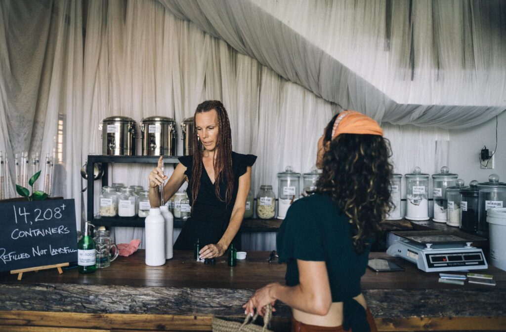 Image of a shop owner behind a counter helping a customer refill reusable shampoo and conditioner bottles. Refillable cosmetics are infinitely more sustainable than throw-away packaging.