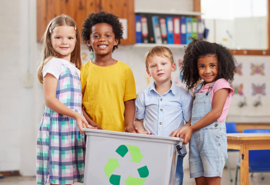 Image of four young smiling children standing in a school classroom holding a recycling container.