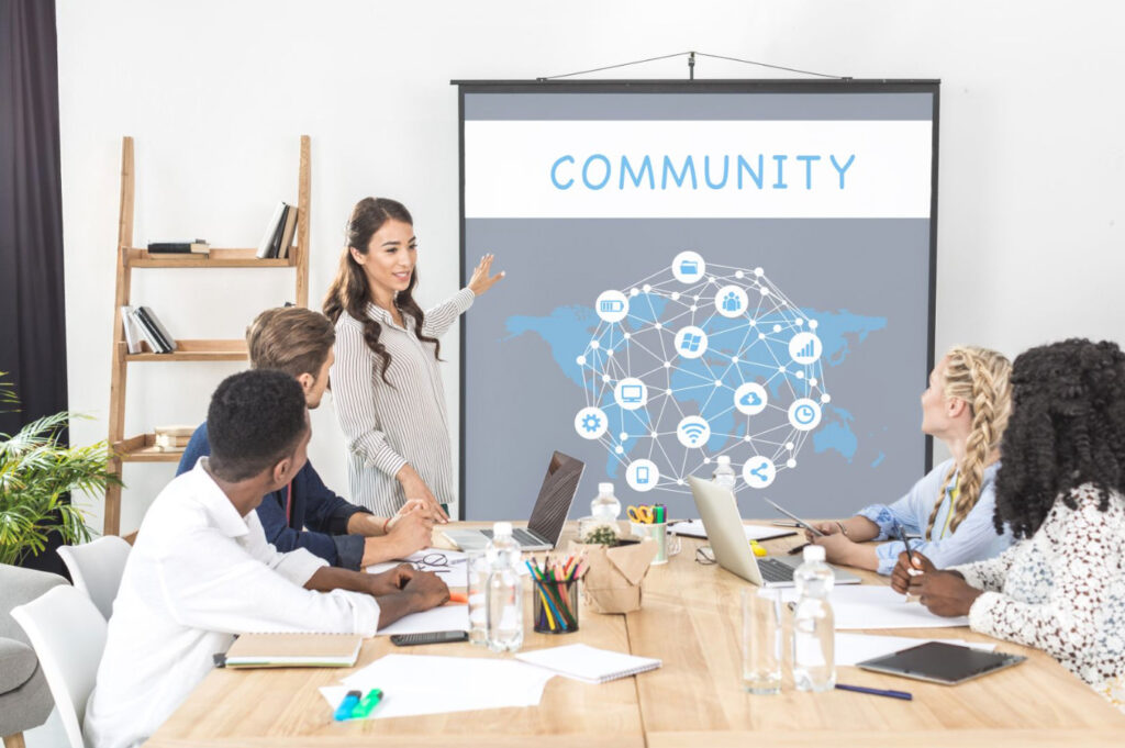 Image of four people sitting at a conference table listening to a speak at the front pointing to a graphic web with the title "Community"