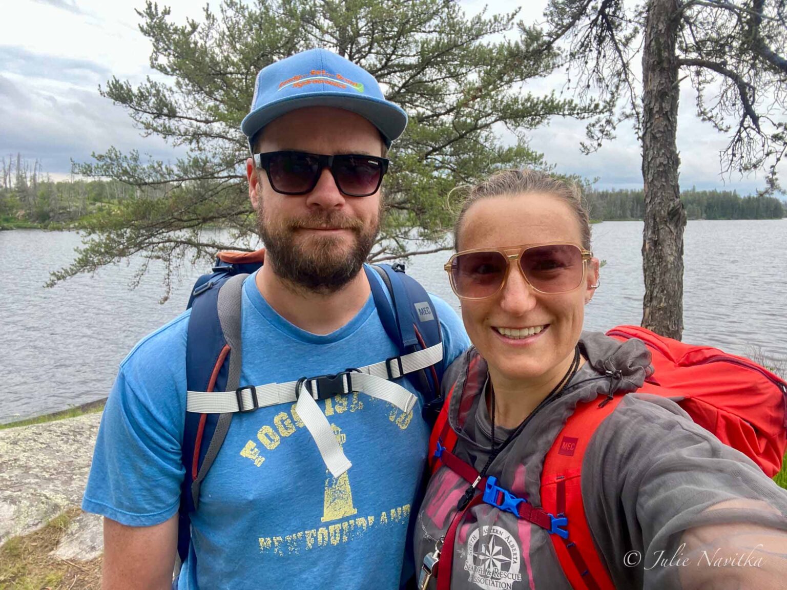 Image of a couple wearing hiking backpacks standing in front of a tree and lake. Being mindful of your impact on the environment is part of sustainable recreation.