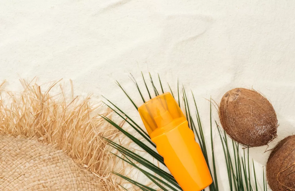Image of a bottle of sunscreen on the sand beside a coconut and grass mat.
