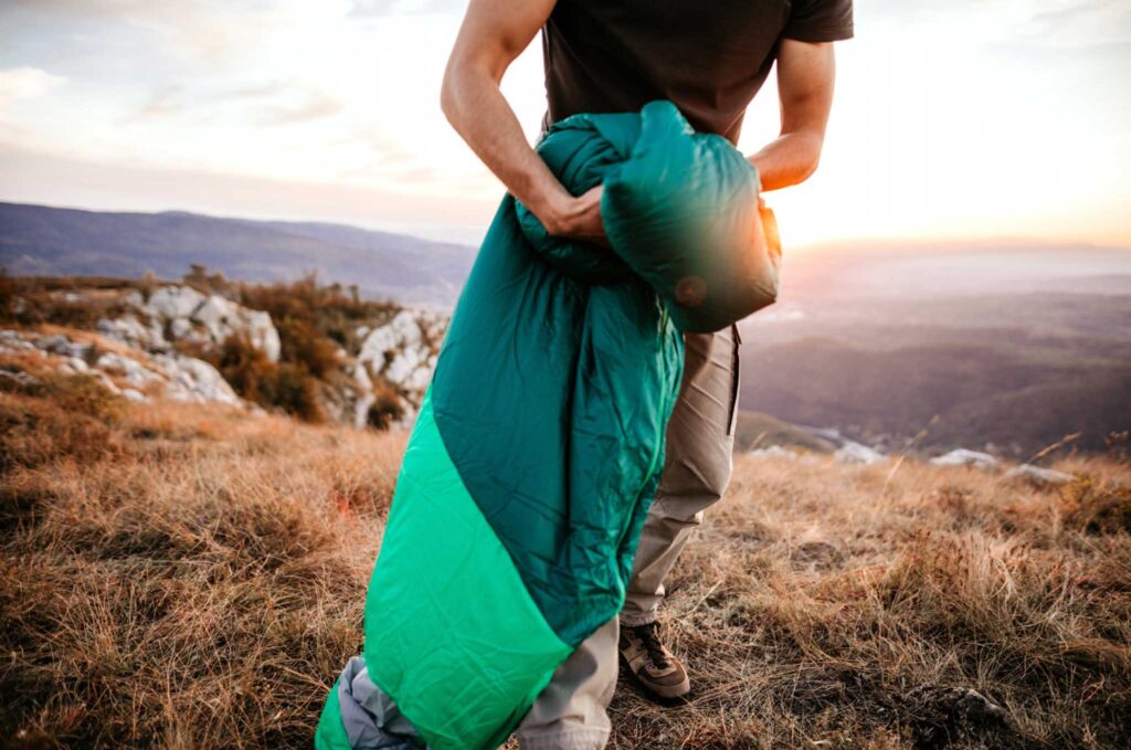 Image of a person packing up a sleeping bag in the great outdoors at sunrise. We must consider the sustainability of all our gear if we want to be true eco-campers.