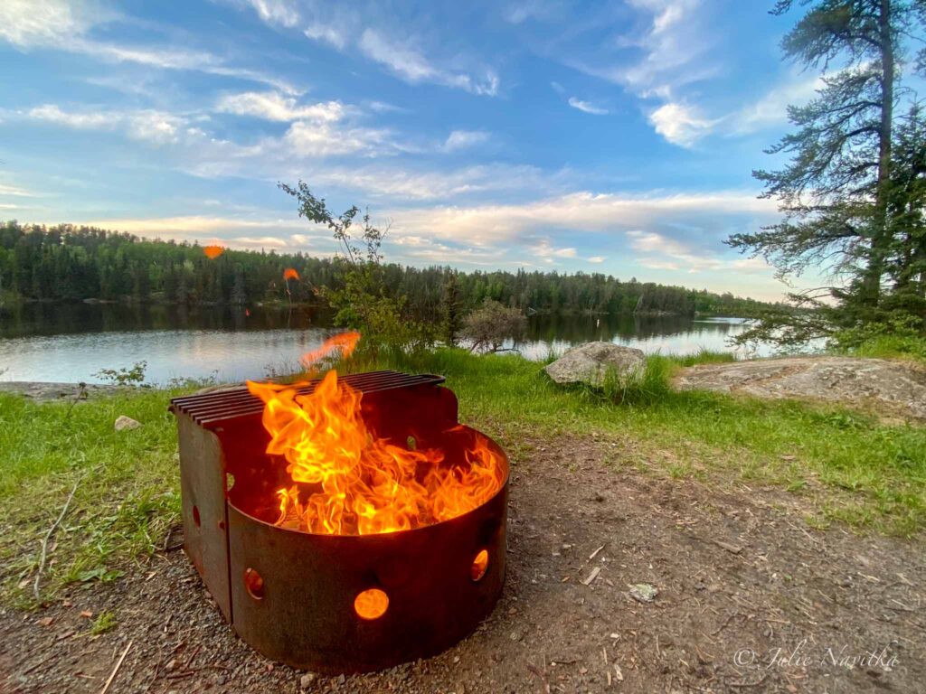 Image of a campfire in a metal pit with lake and blue sky in background. Respecting all fire policies and bans is essential while eco-camping.