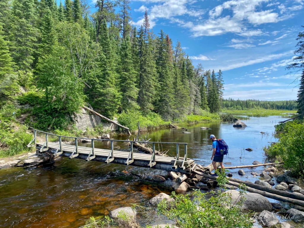 Image of a hiker with backpack crossing a wooden bridge over a small river surrounded by trees on a sunny day. Always leave a plan with someone when hiking to camp in the backcountry.