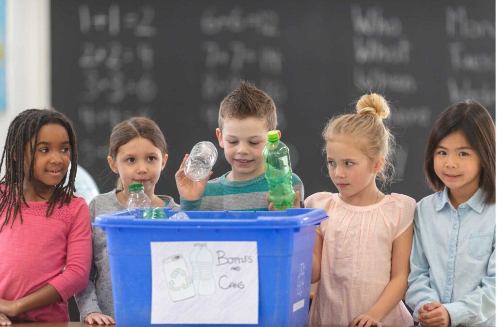 Image of five young students around a recycling bin labelled "bottles and cans". Using the recycle bins properly at school can make for a more sustainable classroom.