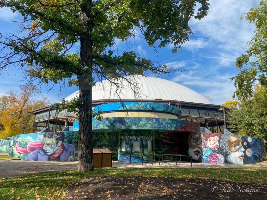 Image of a colorful domed-roof building in a park that reads "Rainbow Stage" above the entry. Building with sustainable materials makes for a more eco-friendly park.