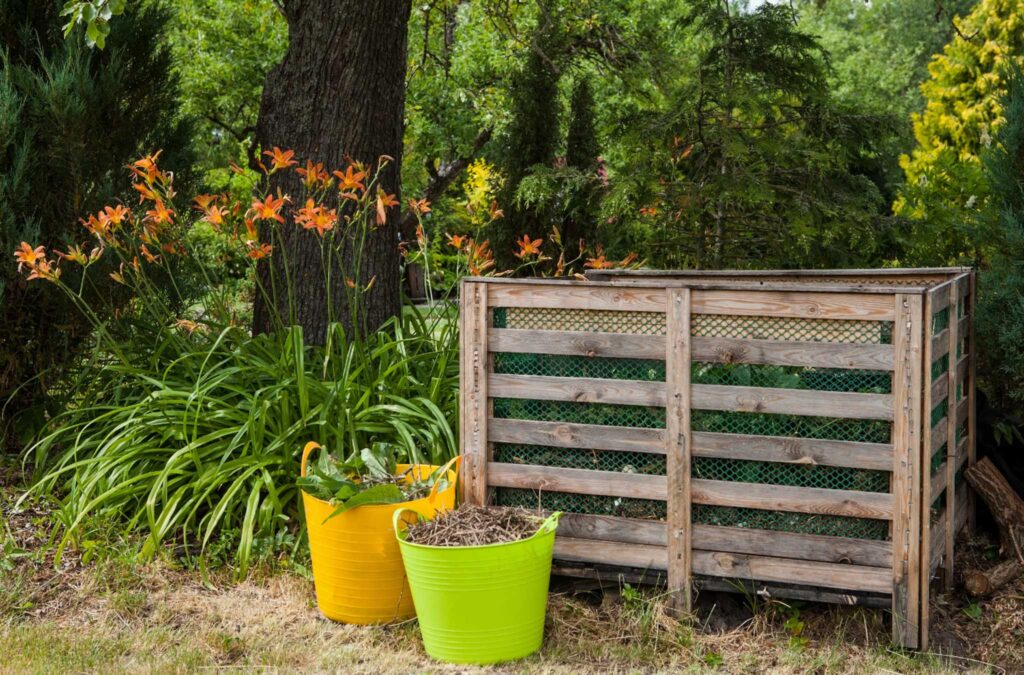 Image of a compost bin made from pallets with some pots in front and day lilies growing beside. Starting a school compost can help your classroom be more eco-friendly.