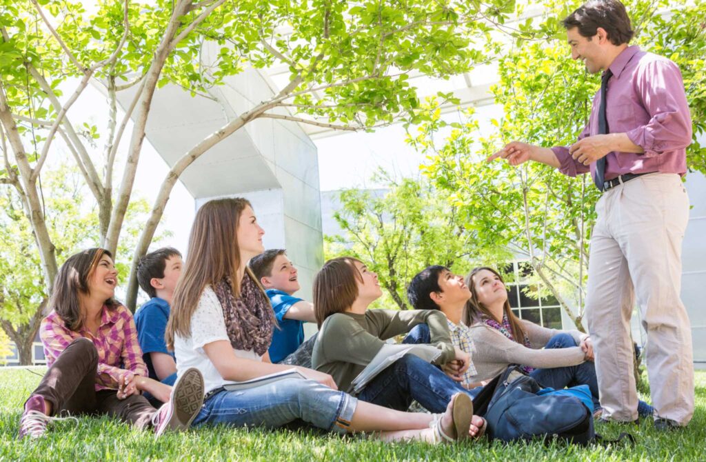 Image of a group of students sitting on the grass outdoors with teacher standing in front interacting with them. Teaching kids about environmental sustainability outdoors is always a great idea.