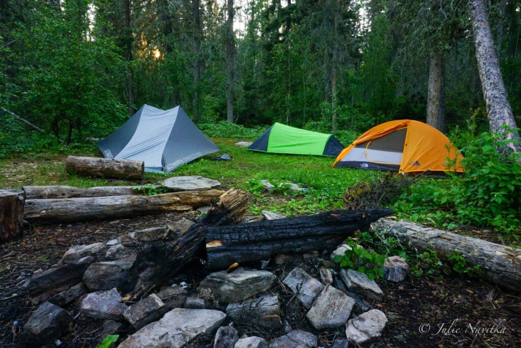 Image of three tents set up in a wooded area with rock fire pit in foreground. There are many sustainable tents to choose from for any camping needs.