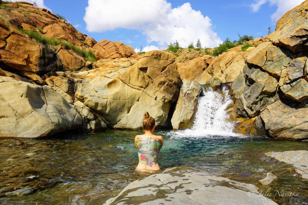 Image of a hiker stopped for a dip to cool off in a small pool at the bottom of a waterfall. Make sure your SPF and bug repellent is all natural if you plan on swimming to protect our waterways while eco-camping.