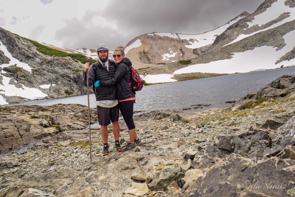 Image of a hiking couple in front of a lake surrounded by mountains with a little bit of snow.