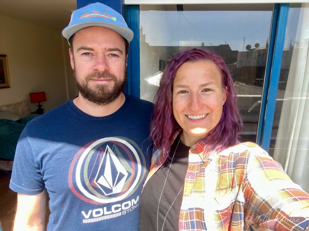 "Selfie" image of the author of the site with their partner standing in the sunshine on an apartment balcony. Slow travelers have less environmental impact than fast-travelling counterparts.