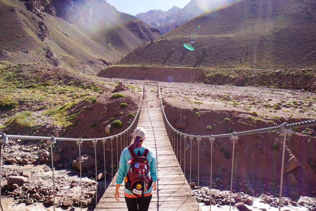 Image of a hiker from behind crossing a wooden suspension bridge strung over a canyon with mountains in background. Ecotourism takes the environment into account.