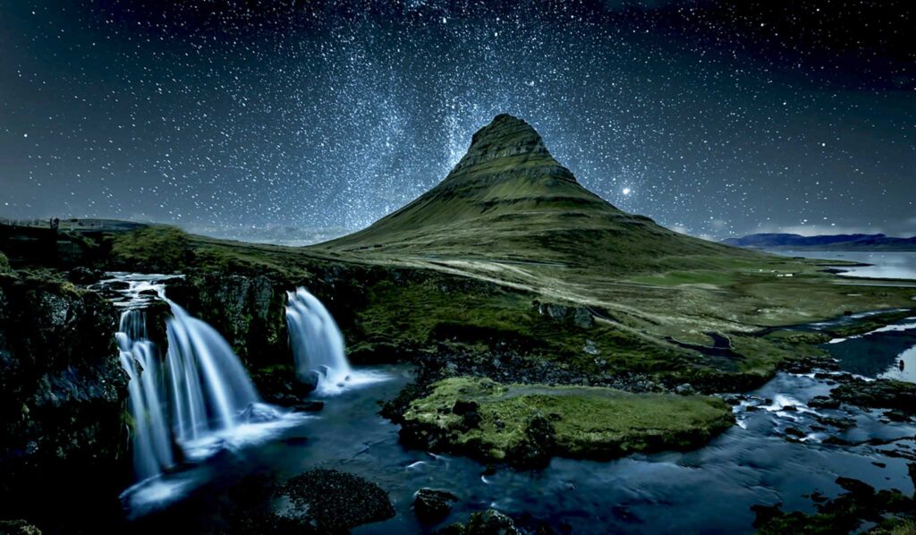 Image of a mountain at night, with waterfalls in the foreground and stars and the milky way illuminating the sky.