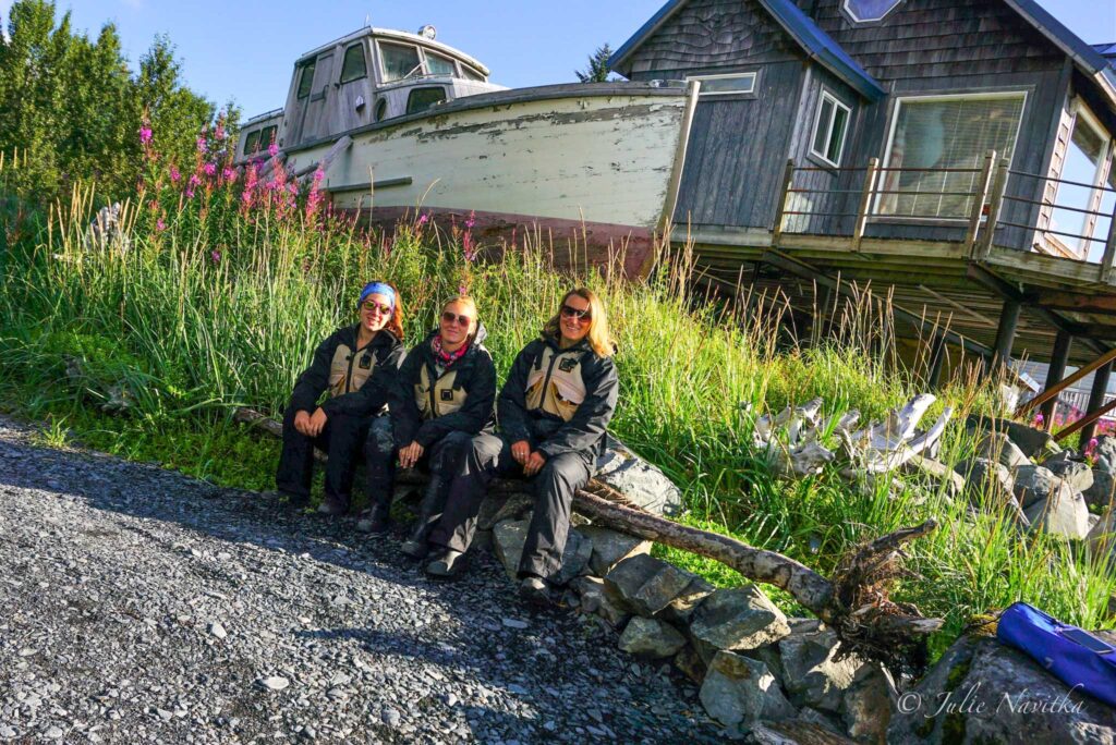 Image of three friends wearing personal flotation devices sitting on a log in front of an old boat waiting to go kayaking.