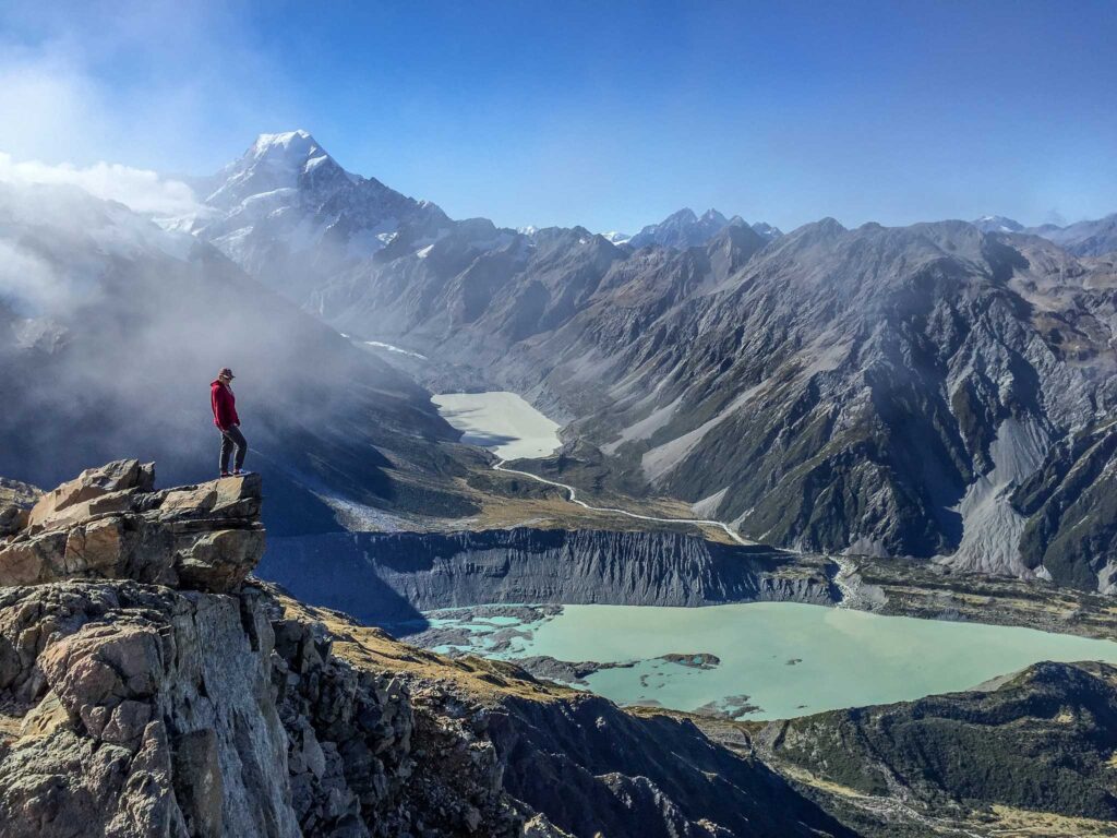 Image of a lone hiker standing on a rocky outcrop high above a chalky turquoise lake with mountains in background. Make sure to follow the leave no trace principles while you practice ecotourism.