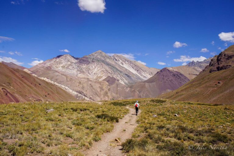 Image of a hiker making their way up a trail toward a mountainscape with a bright blue sky. Slomads should protect the planet by practicing leave no trace principles.