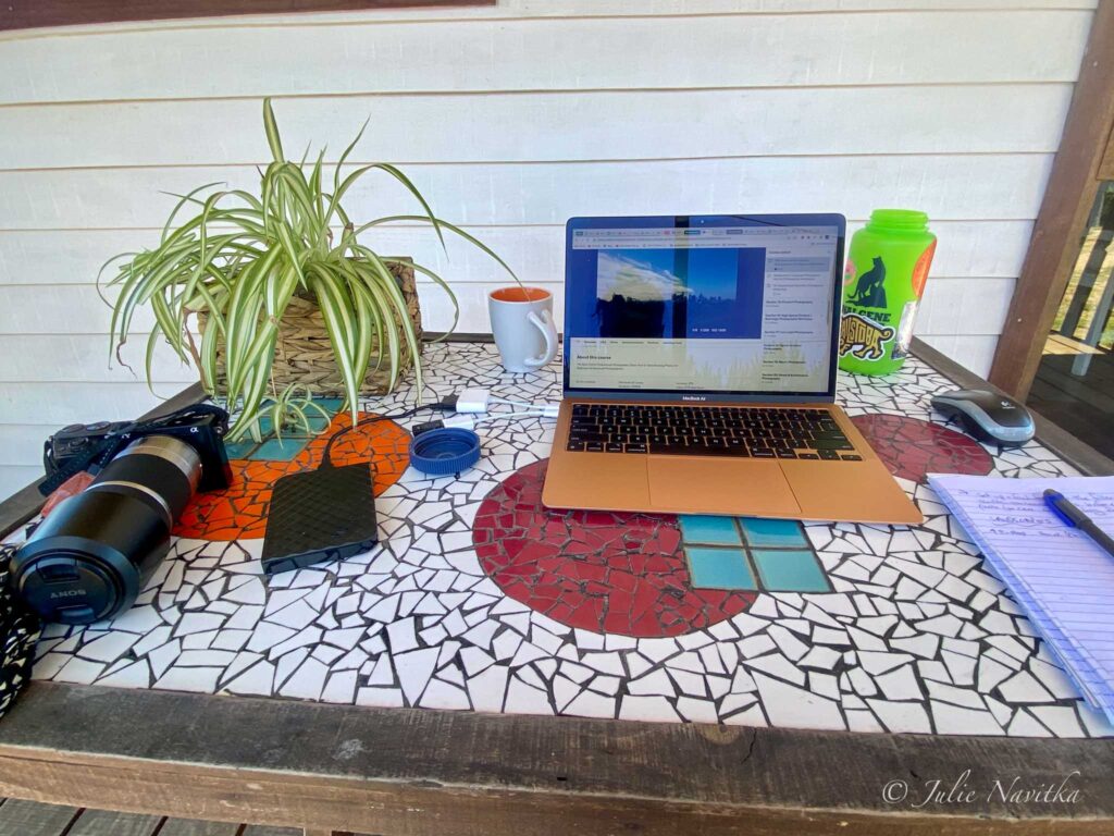 Image of a laptop, camera and other "work" supplies on a mosaic tiled table top outdoors on a porch. Digital nomads can work remotely from anywhere this is wifi.