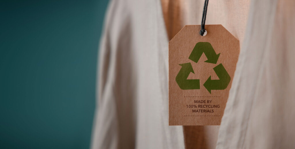 Image of a clothing tag hanging from a cream coloured sweater, made from brown paper with the recycle triangle printed on it.
