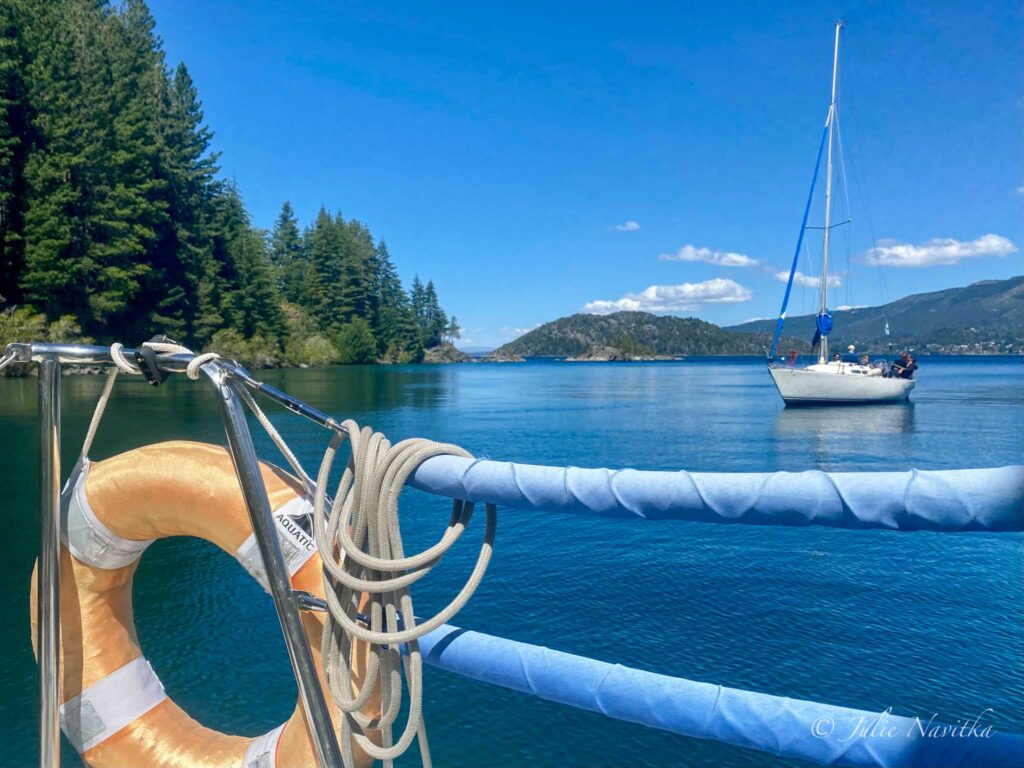 Image taken from a sailboat of another sailboat with life ring in foreground and trees and mountains in background. 