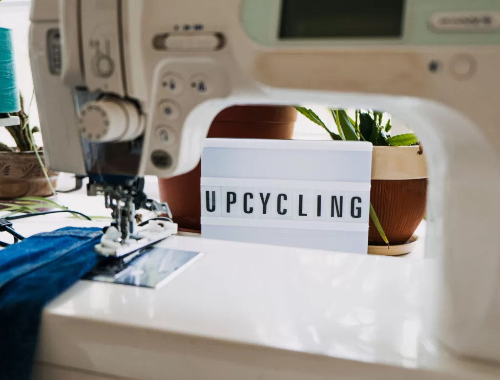 Image taken through a sewing machine of a sign leaning against a plant that says "Upcycling"