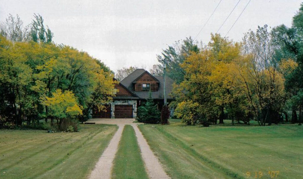 Old image of the front of a home down a long gravel driveway, flanked by grass. A date in the corner reads 2007.
