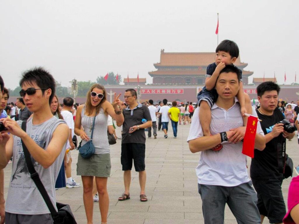 Image of a crowded square in Beijing, China. One tourist looks at the camera and gives a peace sign. What to pack for a year of travel might depend on your destinations.