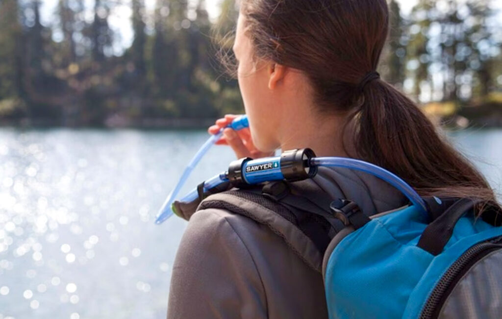 Image of a hiker using the sawyer water filter taken from over her shoulder. This item should definitely be on your "what to pack for a year of travel" list.