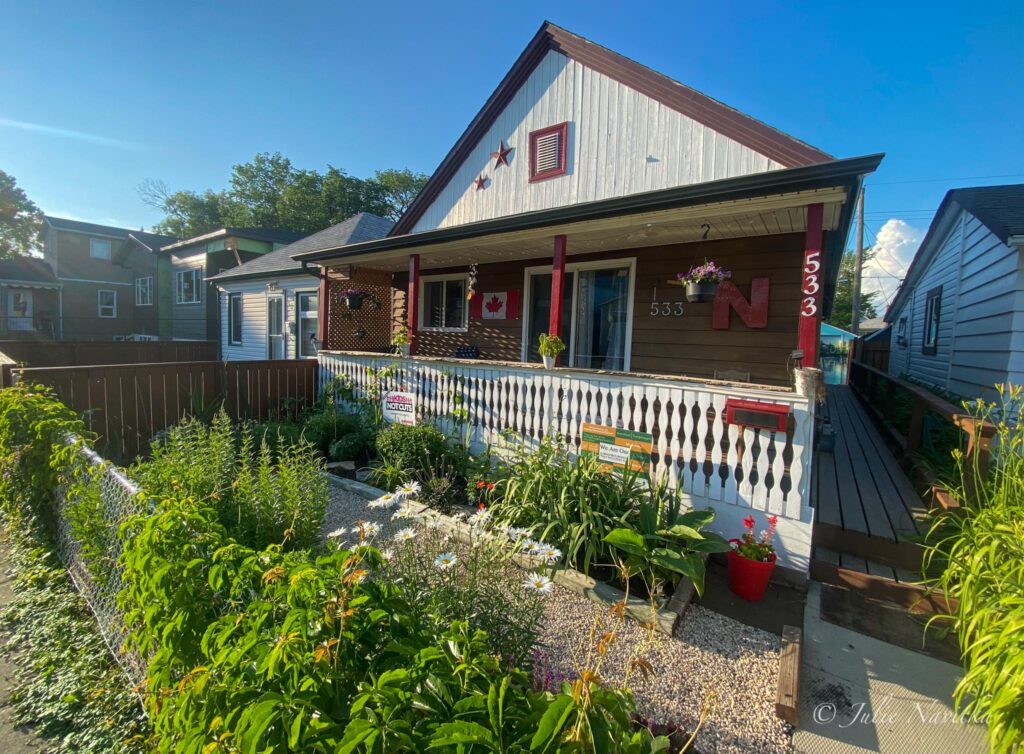 Image of the front of a small home with a grass-free front yard featuring pea gravel, flower beds and vines. Going grass-free in your yard can reduce your water use and carbon footprint.