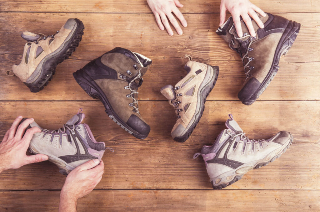 Image from above of a variety of hiking boots on a table with two pairs of hands touching them.